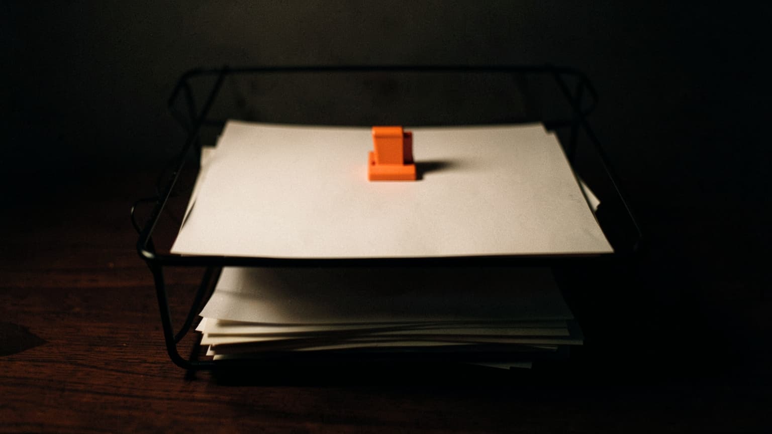 A two-tier letter tray with one finished sheet under an orange clip above an untidy stack of paper
